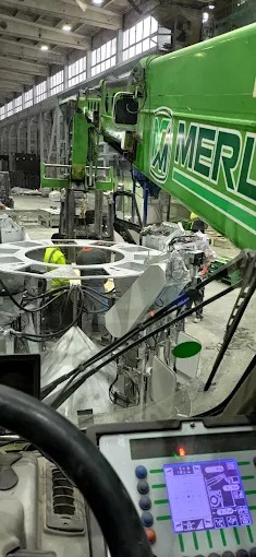 Wnętrze hali: widok z kabiny dźwigu na konstrukcję stalową z pracownikami w żółtych kamizelkach. Dźwig marki Merlo w trakcie pracy.