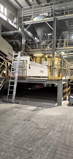 Wnętrze hali produkcyjnej z metalową konstrukcją wsporczą, schodami i drabiną. Widoczna maszyna z napisem „HAVER”.