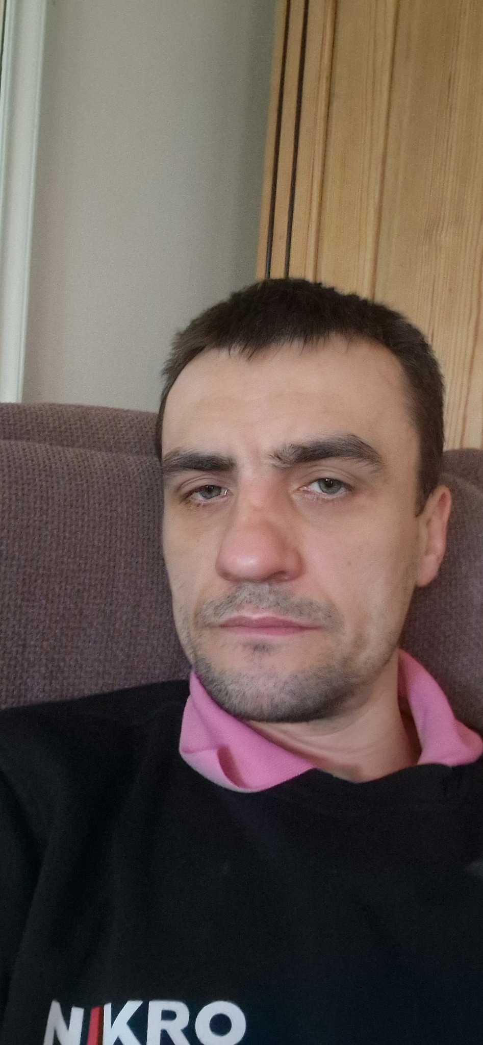Portret mężczyzny z krótkimi włosami, ubranego w czarną bluzę z widocznym logo 'NIKRO' i różową koszulę, siedzącego na brązowym fotelu na tle jasnej ściany.