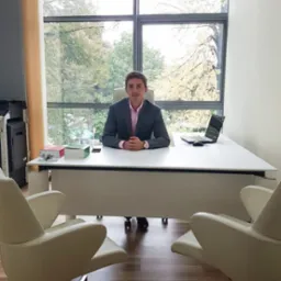 Młody prawnik w szarej marynarce siedzi za biurkiem w nowoczesnym biurze z dużym oknem. Na biurku laptop i dokumenty, z przodu dwa fotele dla klientów.