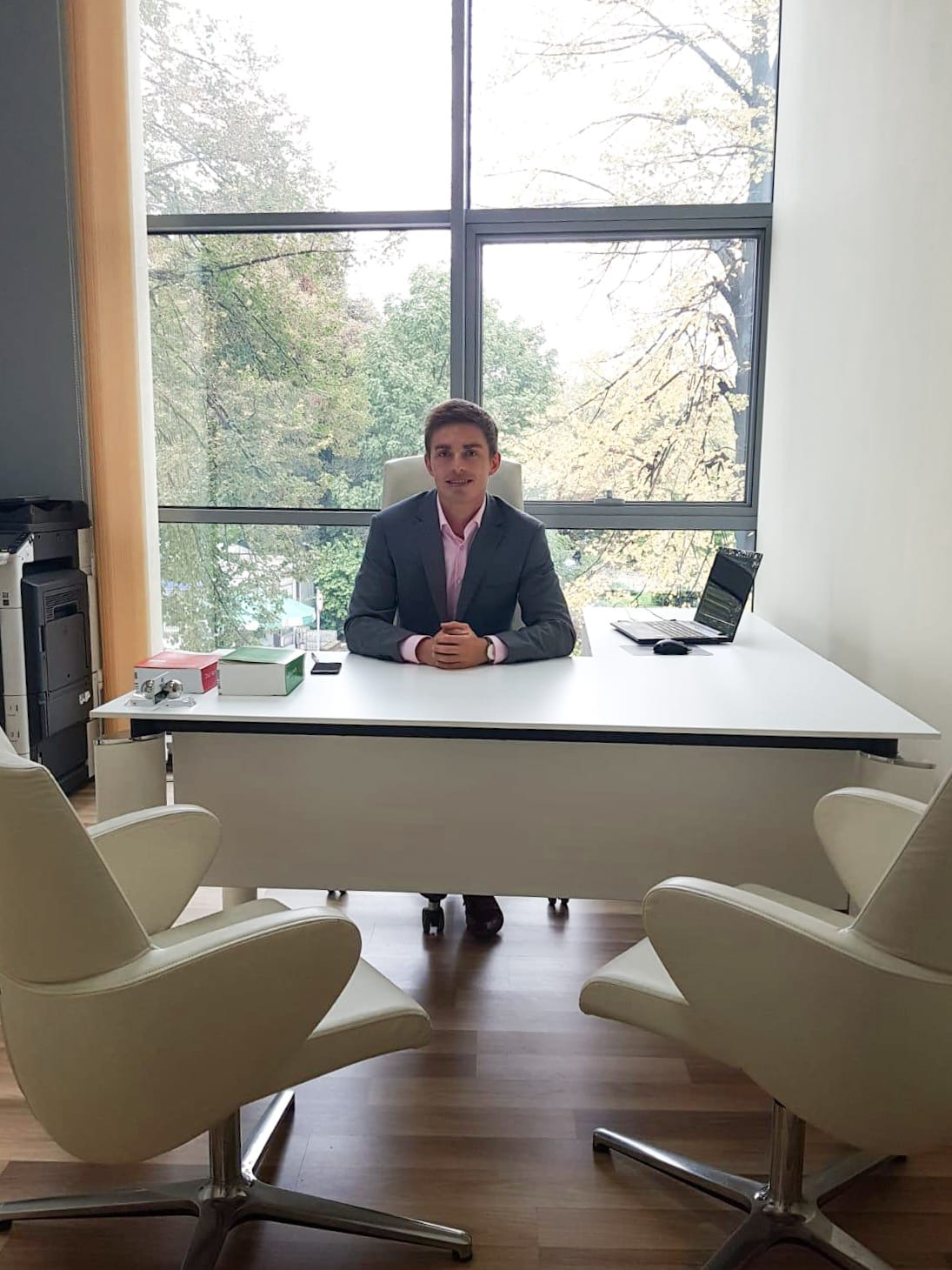 Młody prawnik w szarej marynarce siedzi za biurkiem w nowoczesnym biurze z dużym oknem. Na biurku laptop i dokumenty, z przodu dwa fotele dla klientów.