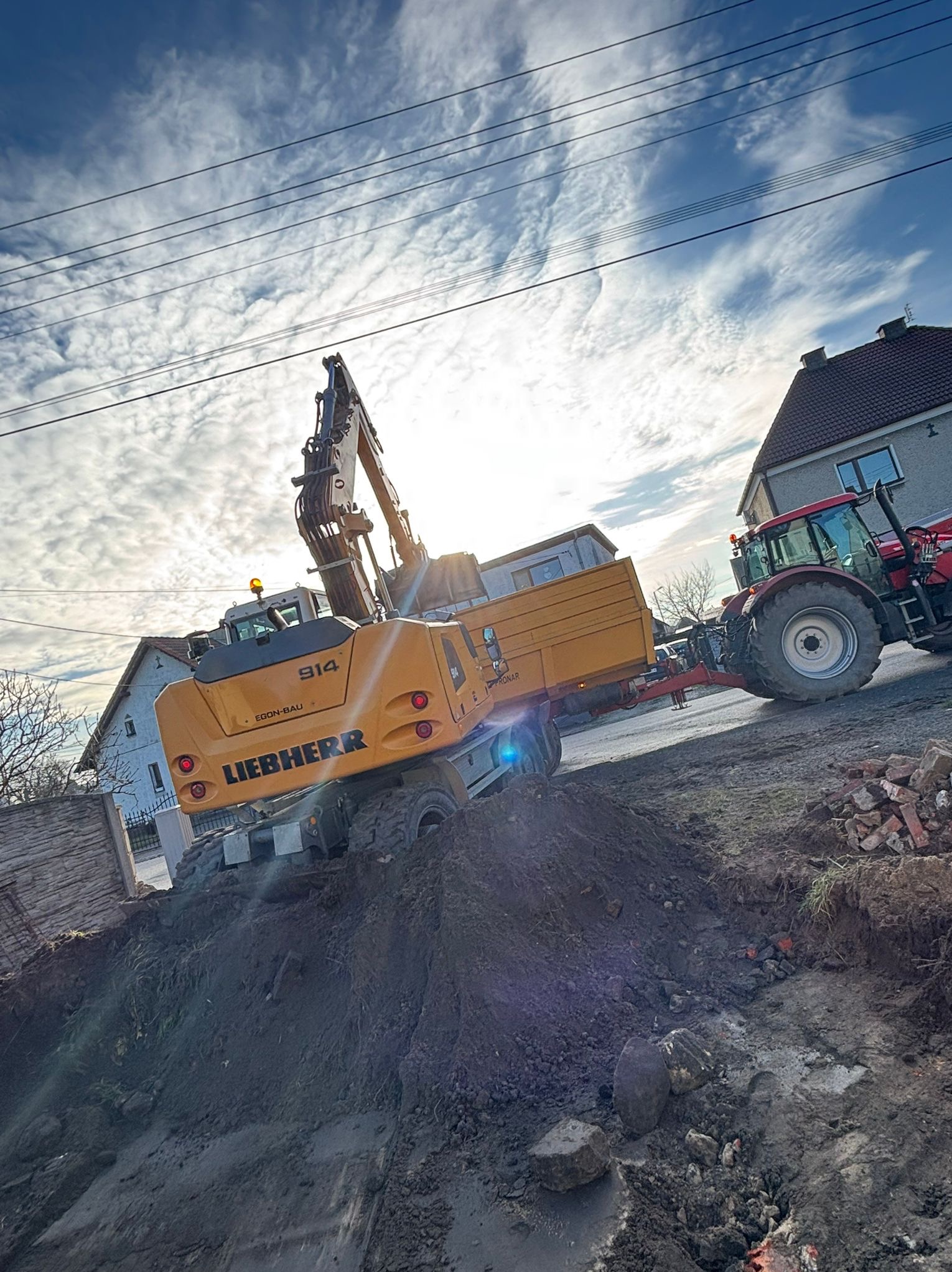 Żółta koparka Liebherr 914 na nierównym terenie z wykopem, w tle dom i traktor, pochmurne niebo. Ujęcie z dołu.