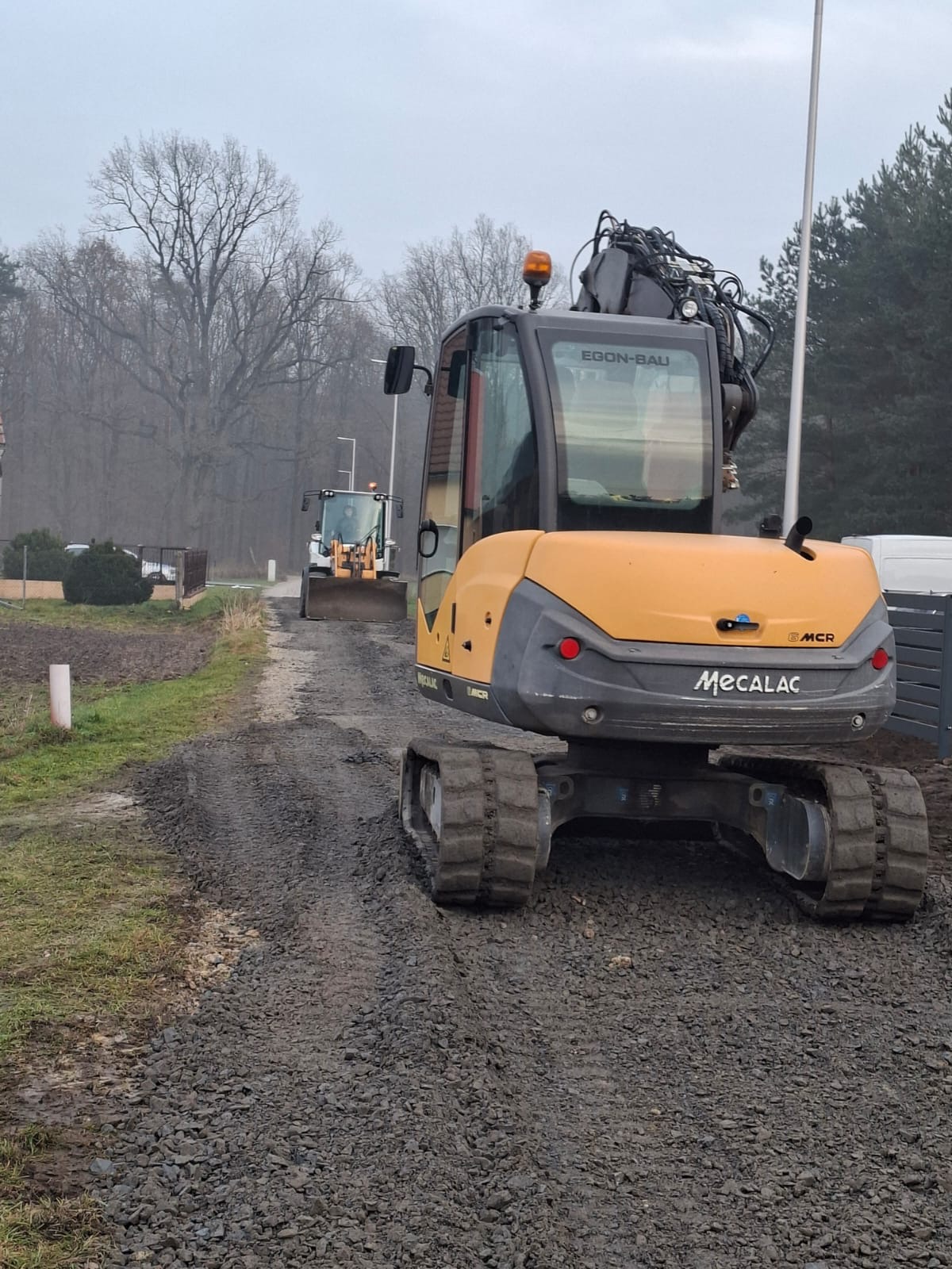 Żółta koparka Mecalac na żwirowej drodze, w tle biała ładowarka. Prace ziemne w terenie zabudowanym, pochmurny dzień.