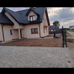 Nowo wybudowany dom z ułożoną kostką brukową na podjeździe i przygotowanym terenem pod przyszły ogród. Osoba w oddali pracuje nad wyrównaniem ziemi.