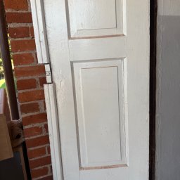 Sztuka ogrodu - Białe, odnowione drzwi z panelami, uchwycone pod kątem, z widoczną czerwoną cegłą w tle. Widoczne ślady farby i niedoskonałości.