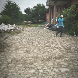 Sztuka ogrodu - Mężczyzna w jasnej koszulce idzie po kamiennej drodze obok sterty gruzu i worków, prawdopodobnie po pracach porządkowych na posesji.