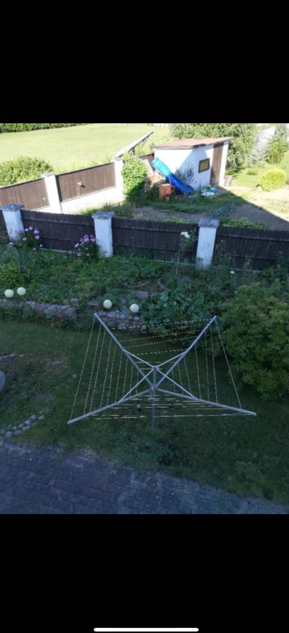 Widok z góry na ogród z ziemią, kwiatami, ogrodzeniem i szopą. Na pierwszym planie rozkładana suszarka do ubrań.