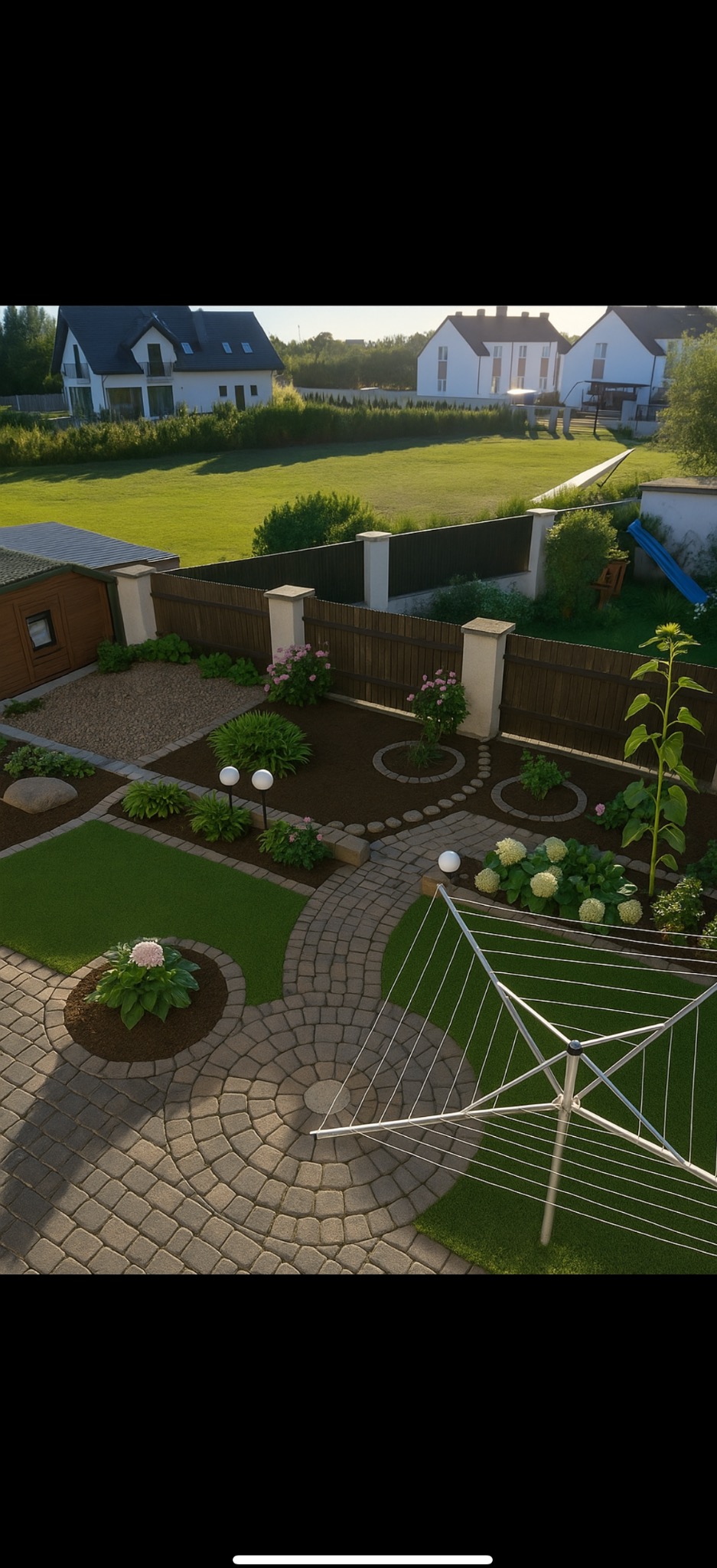 Widok z góry na zadbany ogród z okrągłym brukowanym patio, rabatami kwiatowymi i równo przystrzyżonym trawnikiem. W tle domy i zielona przestrzeń.