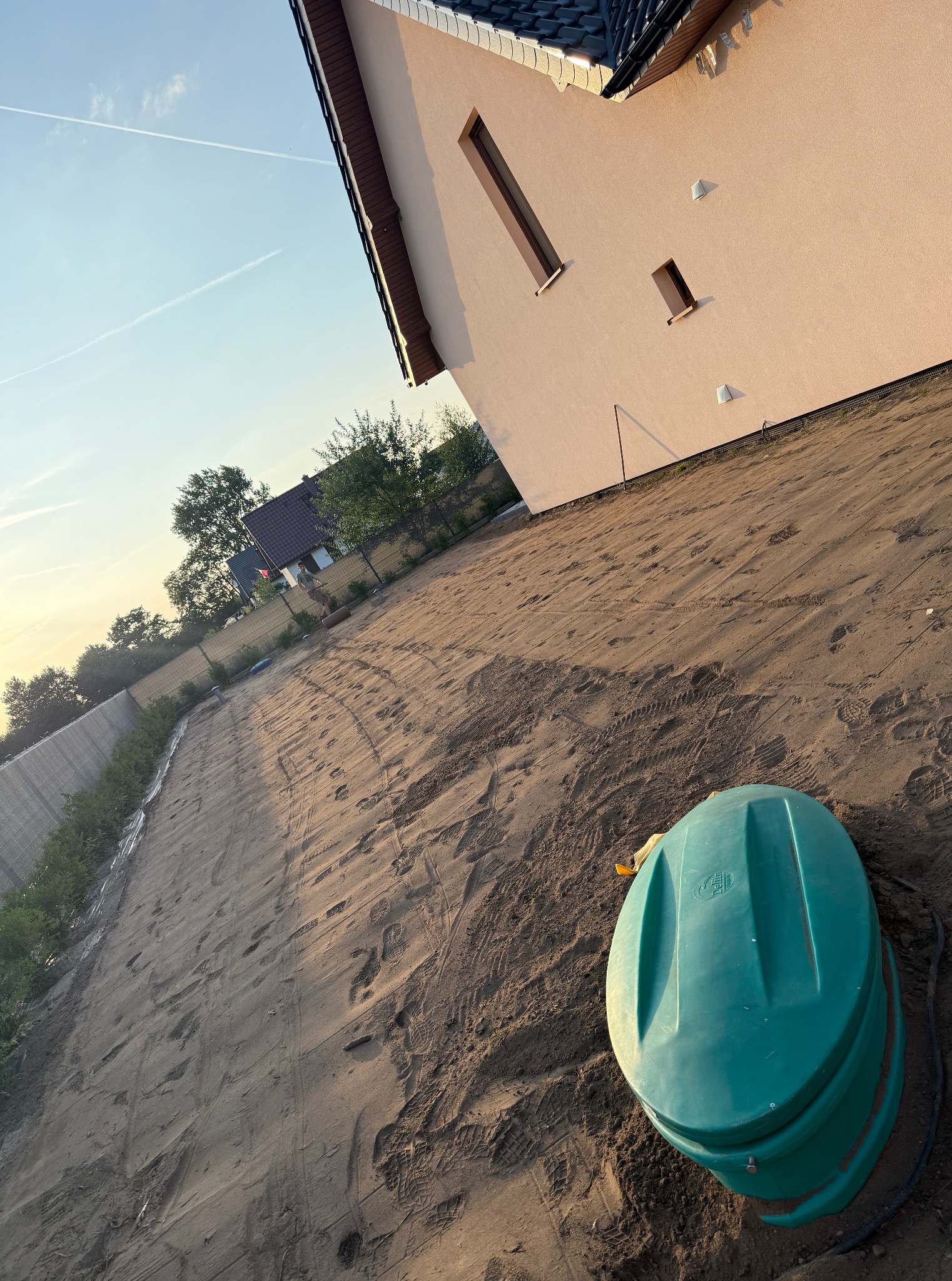 Przygotowany teren pod przyszły ogród z widocznym zbiornikiem na deszczówkę. Ślady opon na piasku sugerują wyrównywanie terenu. Dom z jasną elewacją w tle.
