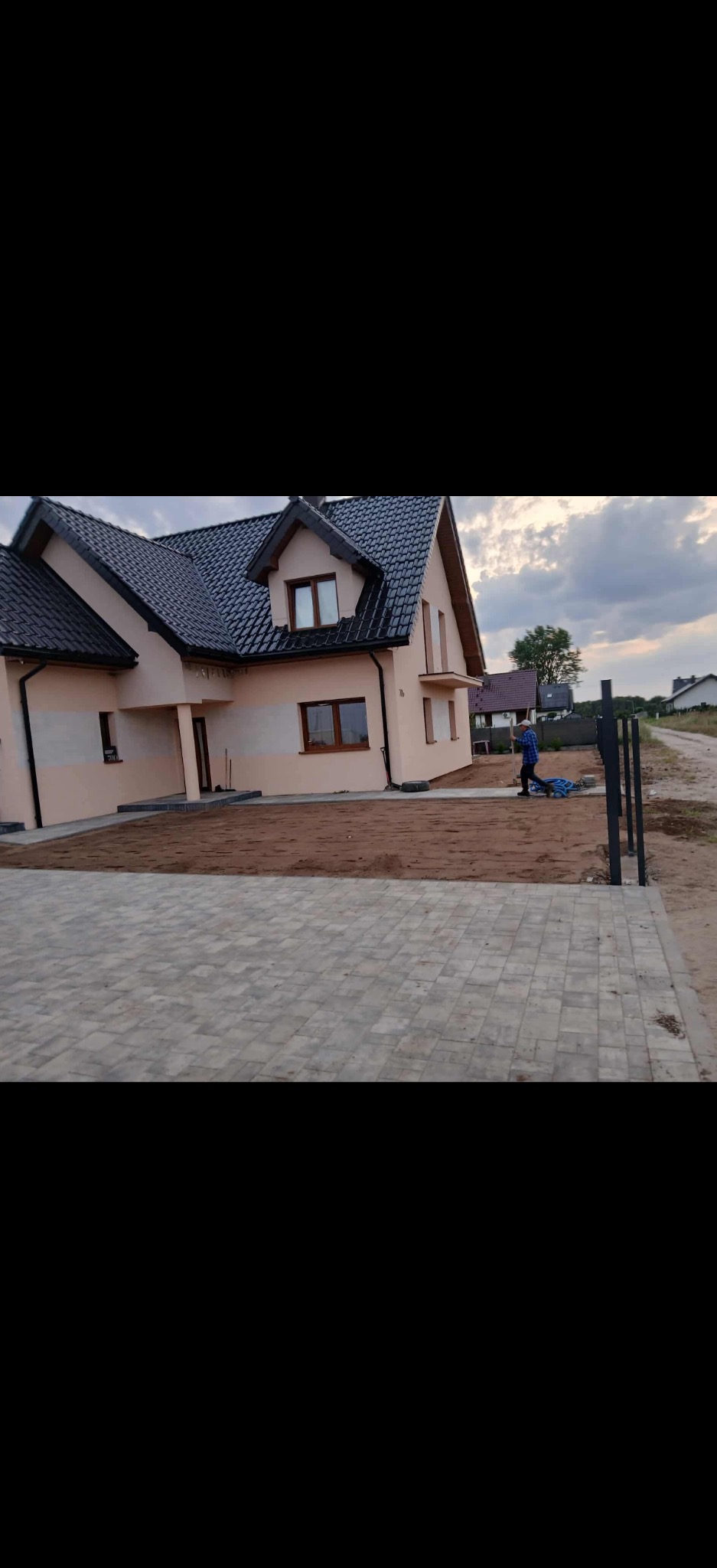 Nowo wybudowany dom z ułożoną kostką brukową na podjeździe i przygotowanym terenem pod przyszły ogród. Osoba w oddali pracuje nad wyrównaniem ziemi.