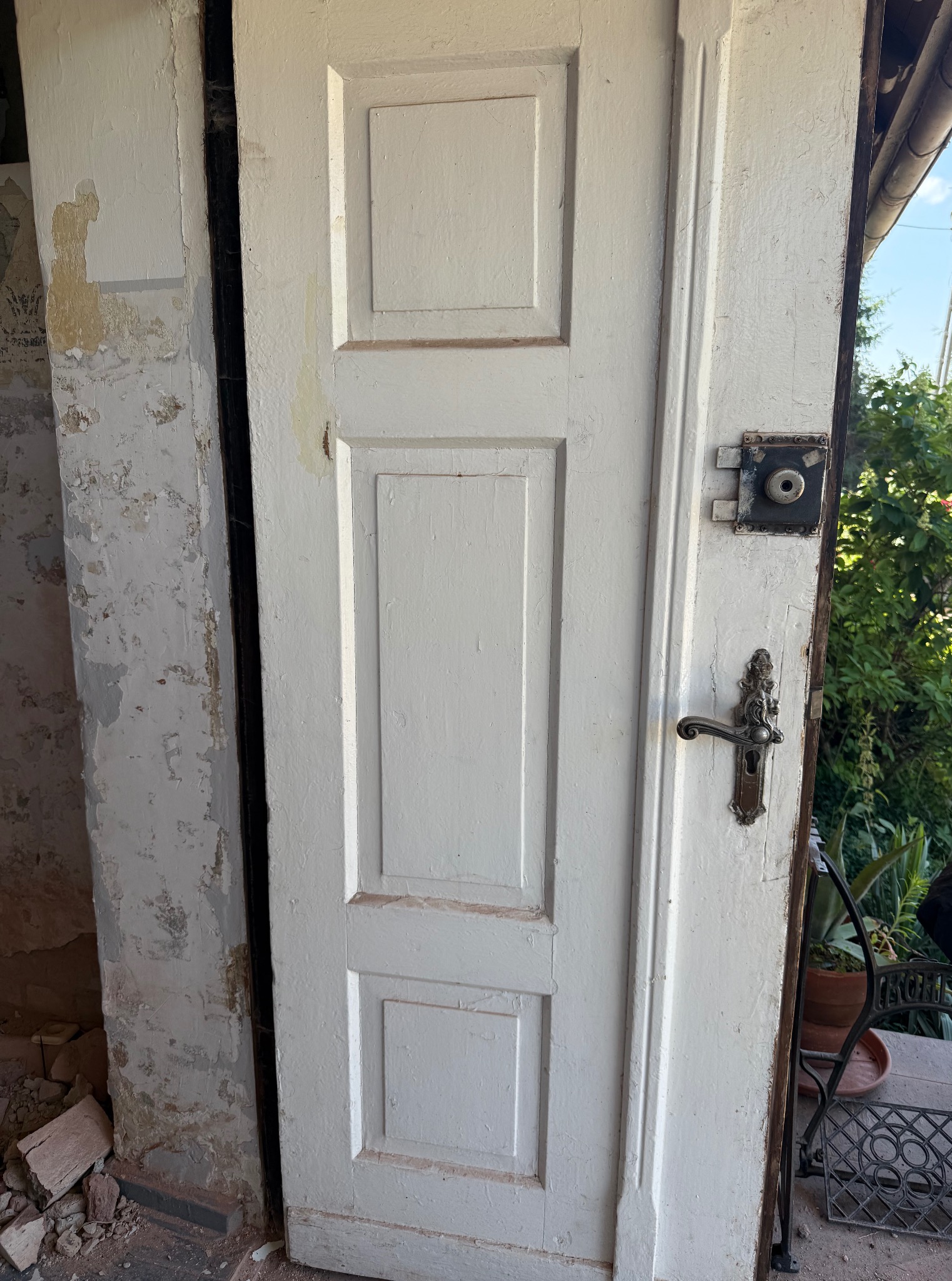 Otwarta, drewniana, biała, skrzydłowa brama z ozdobną klamką, widoczna na tle zieleni ogrodu. Fragment ściany z odpadającym tynkiem po lewej stronie.