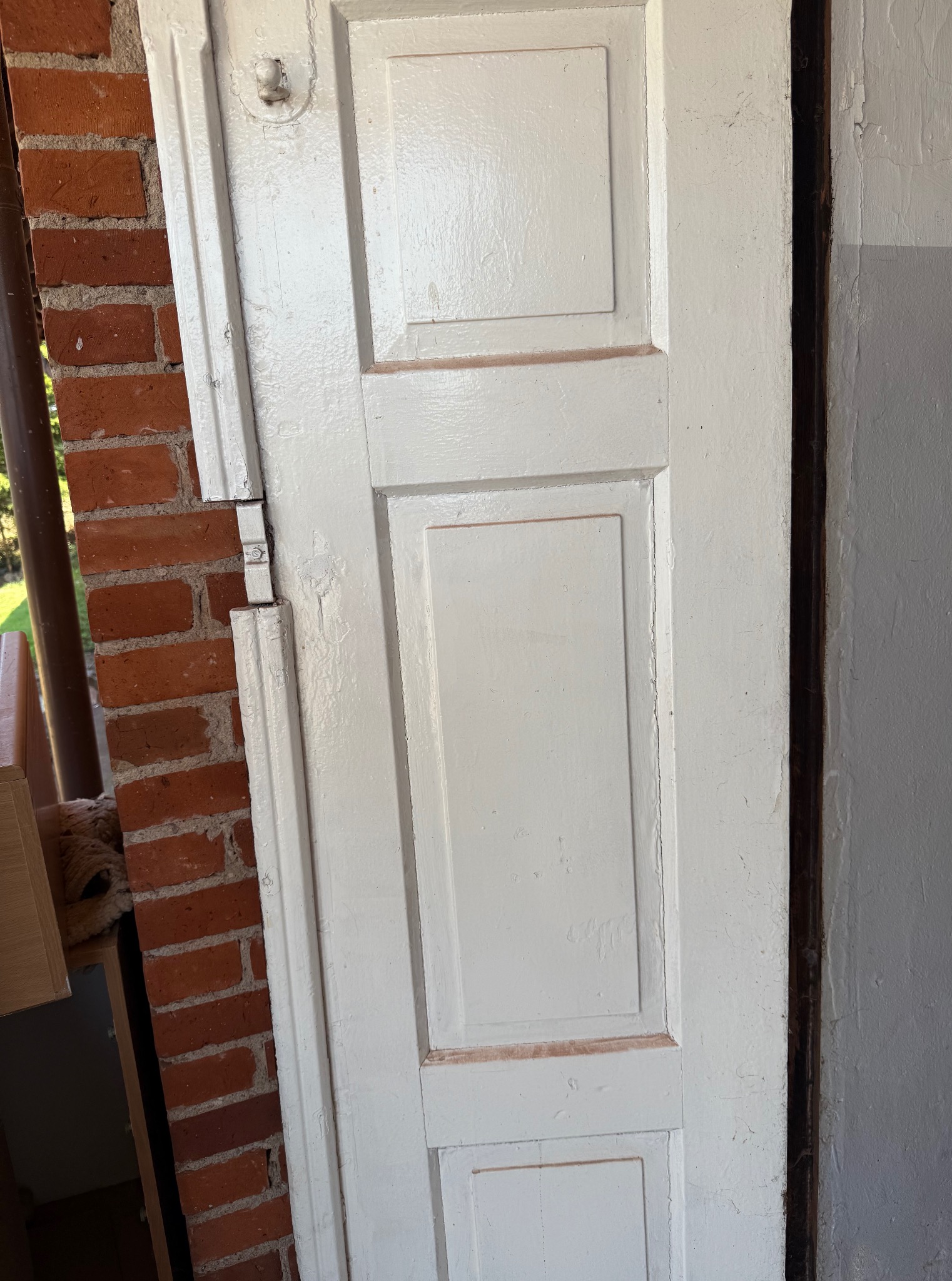 Białe, odnowione drzwi z panelami, uchwycone pod kątem, z widoczną czerwoną cegłą w tle. Widoczne ślady farby i niedoskonałości.
