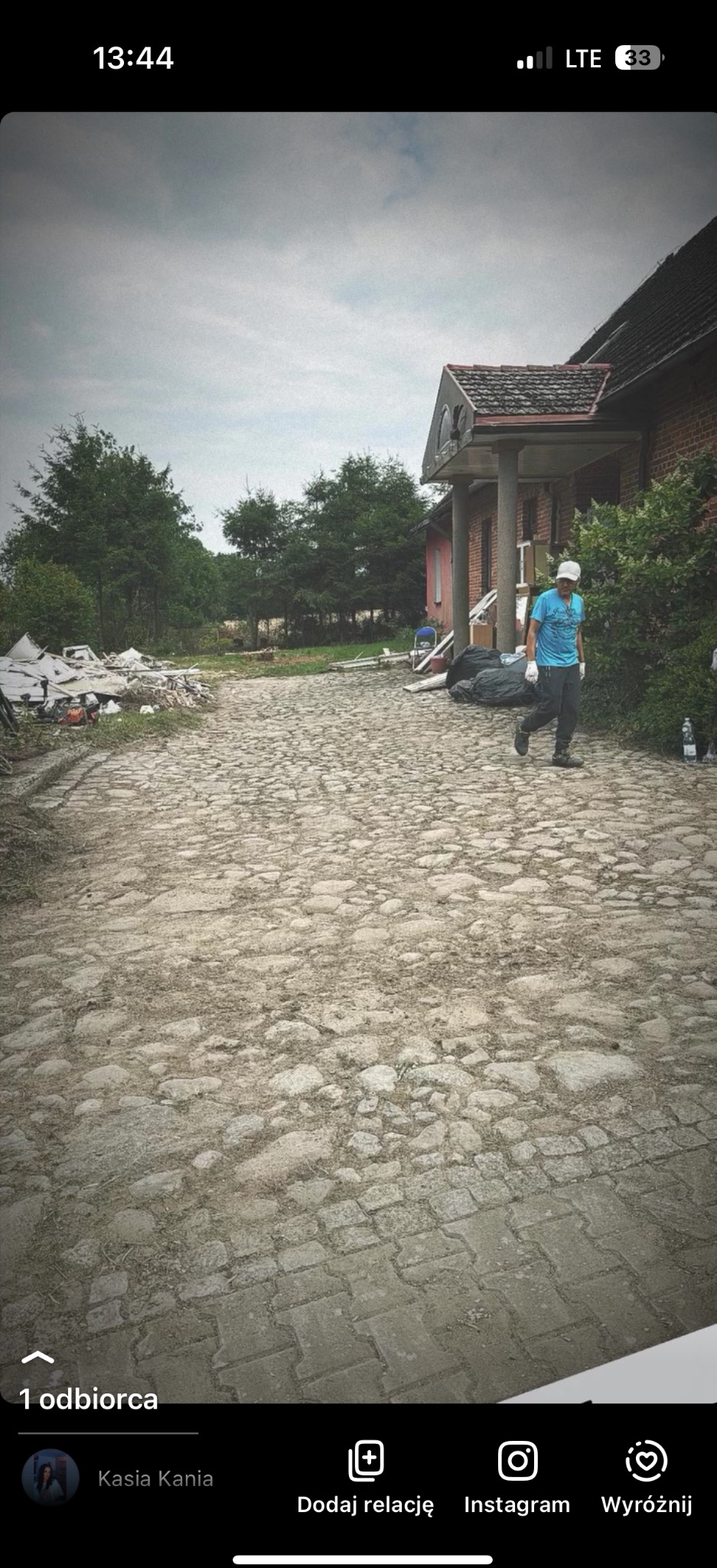 Mężczyzna w jasnej koszulce idzie po kamiennej drodze obok sterty gruzu i worków, prawdopodobnie po pracach porządkowych na posesji.