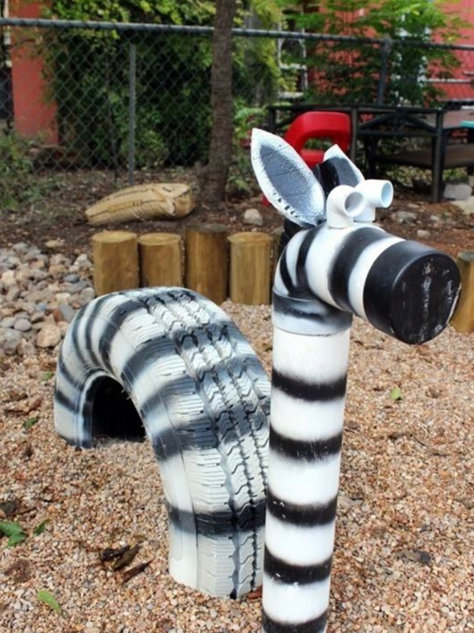 Oryginalna dekoracja ogrodu: zebra wykonana z pomalowanej opony i rur PCV, ustawiona na żwirowym podłożu, z drewnianymi elementami w tle.