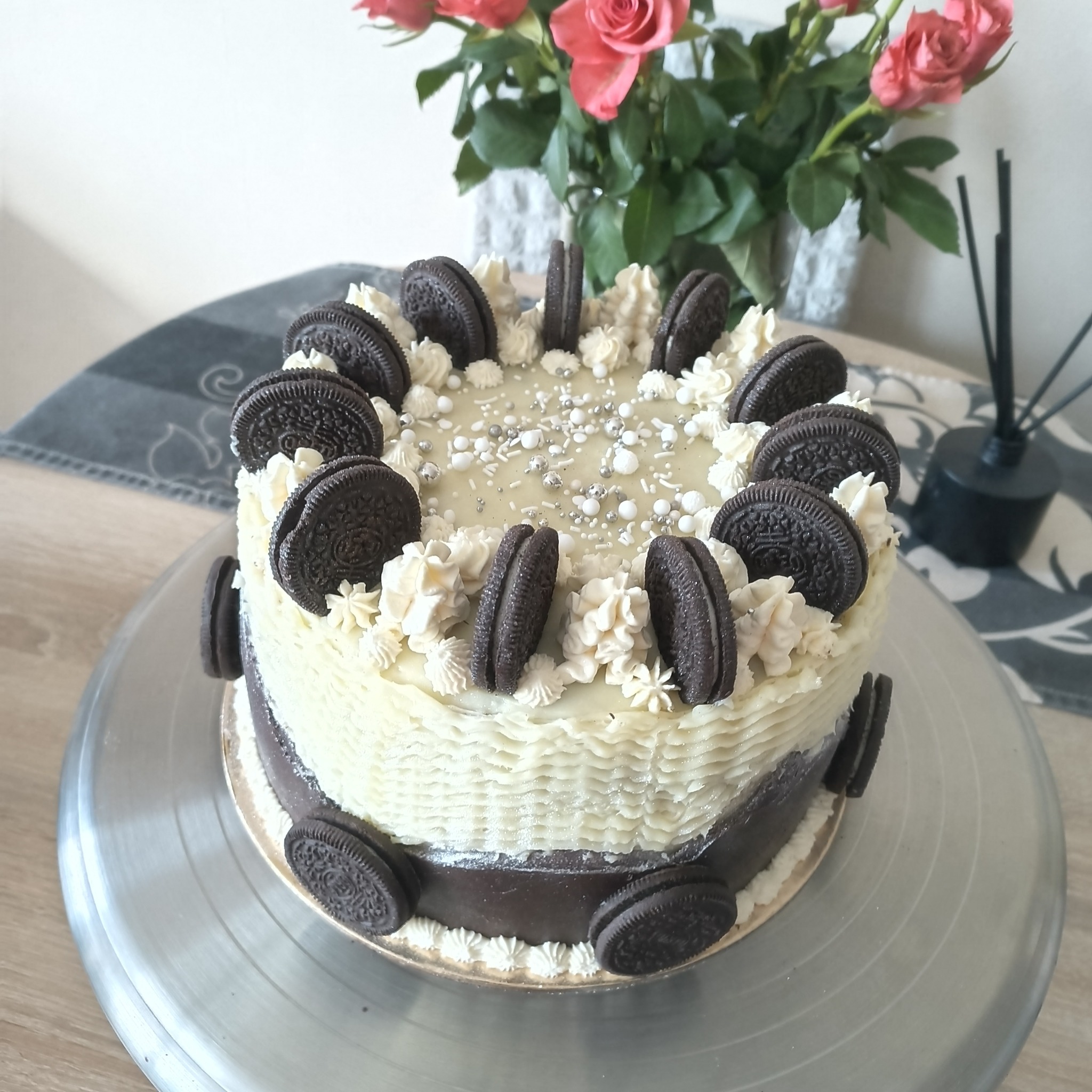 Tort Oreo z kremem, posypką i różami w tle, na srebrnej paterze. Krem w kolorze złamanej bieli, z widoczną teksturą. Dekoracja z ciasteczek Oreo.
