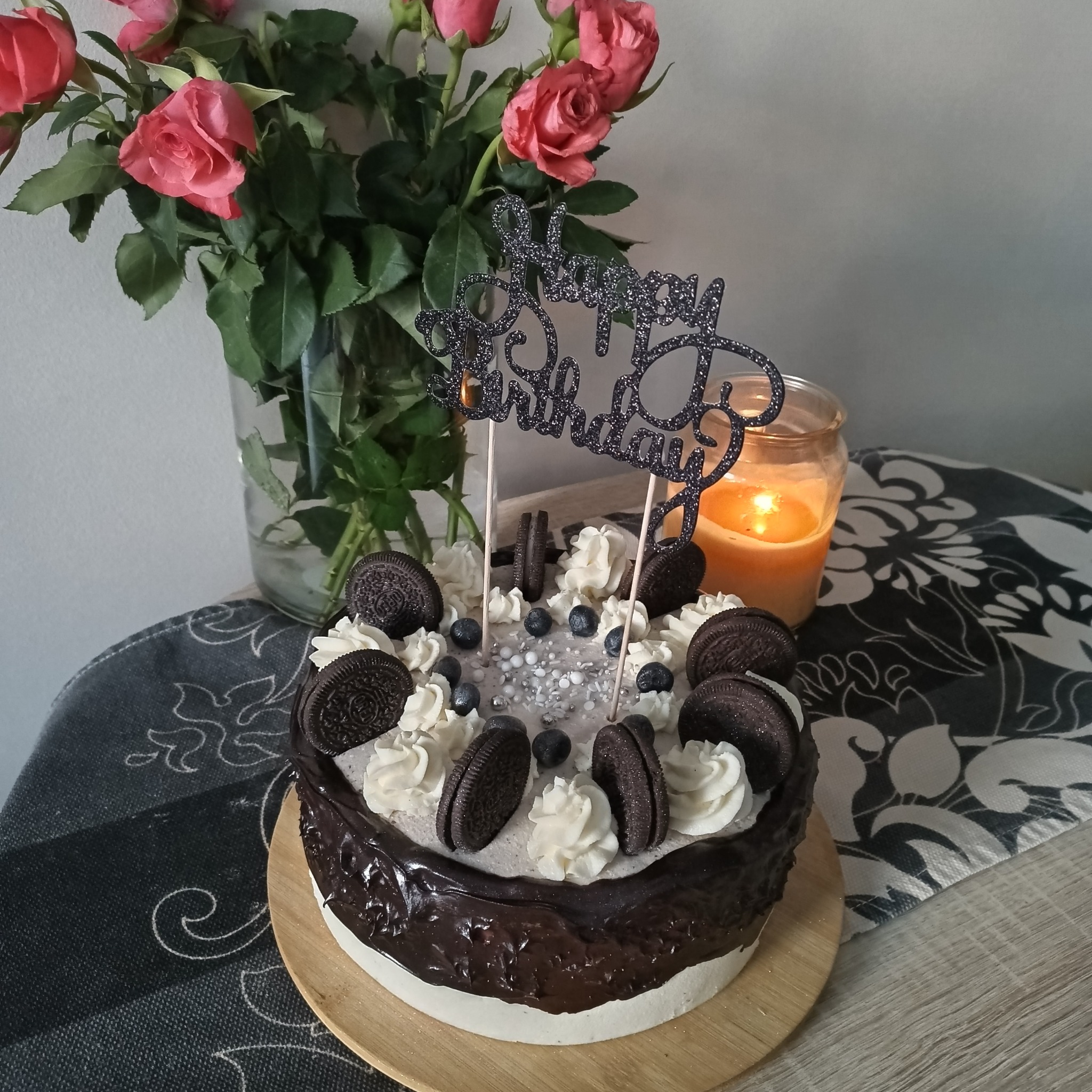 Tort urodzinowy z dekoracją Oreo, kremem i borówkami, na drewnianej podstawie, w tle bukiet róż i świeczka. Słodki deser na specjalną okazję.