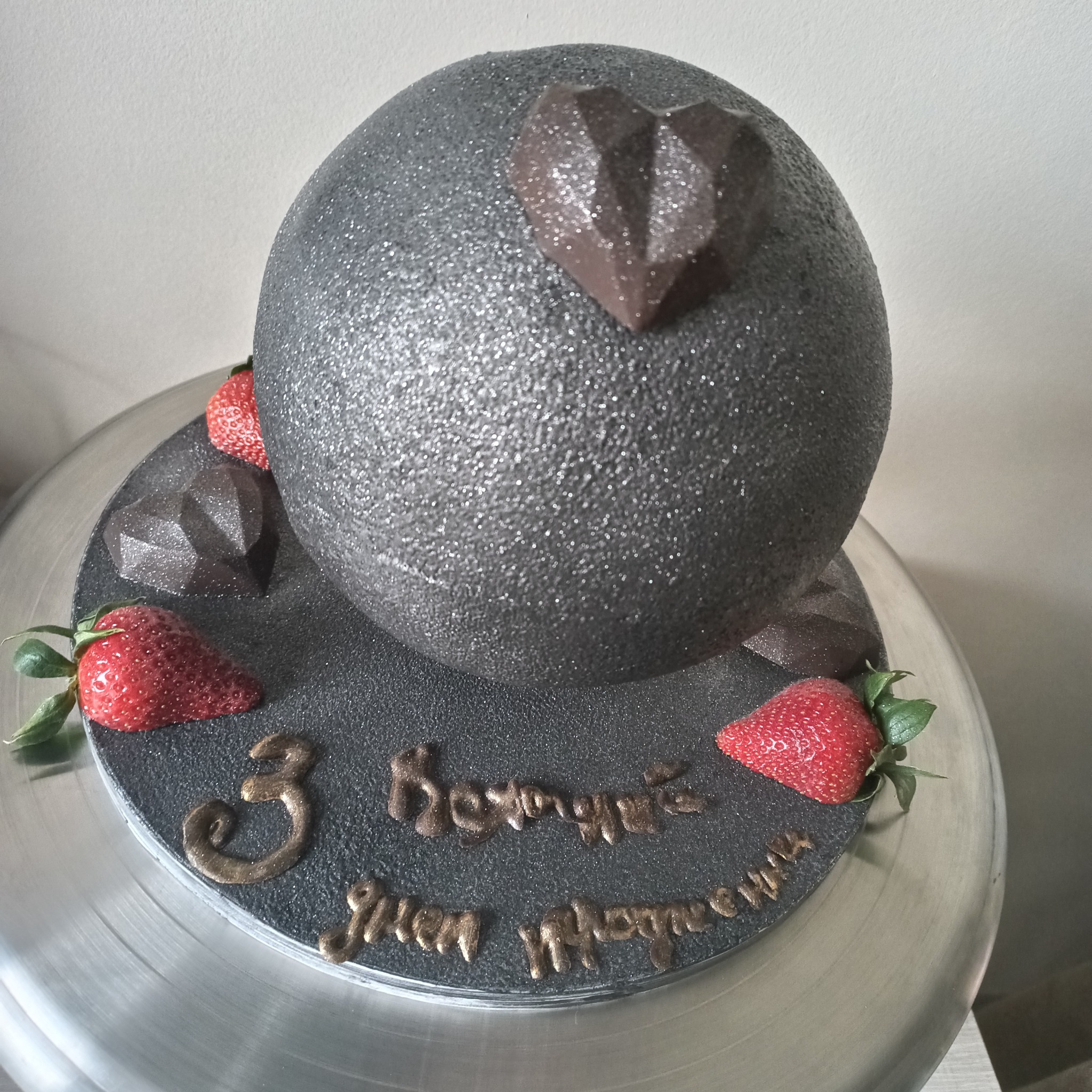 Szary, błyszczący tort w kształcie kuli z czekoladowym diamentem, truskawkami i napisem '3 Koronacie Irenie i Małgorzacie' na srebrnej paterze.