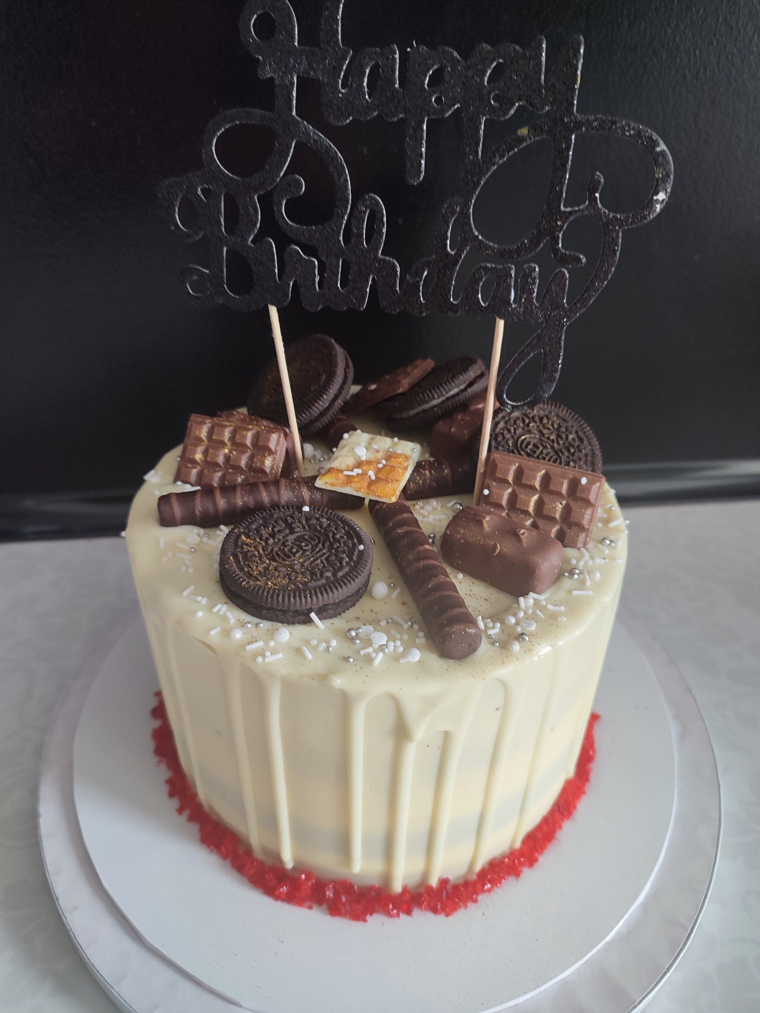 Tort urodzinowy z polewą z białej czekolady, dekorowany ciasteczkami Oreo, batonikami i posypką, zwieńczony czarnym napisem 'Happy Birthday'.