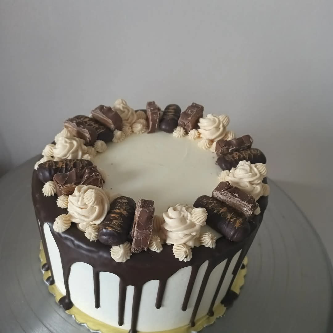 Tort z białym kremem, polewą czekoladową i dekoracjami z batoników, ustawiony na srebrnej paterze. Widoczne detale kremowych rozetek i spływającej polewy.