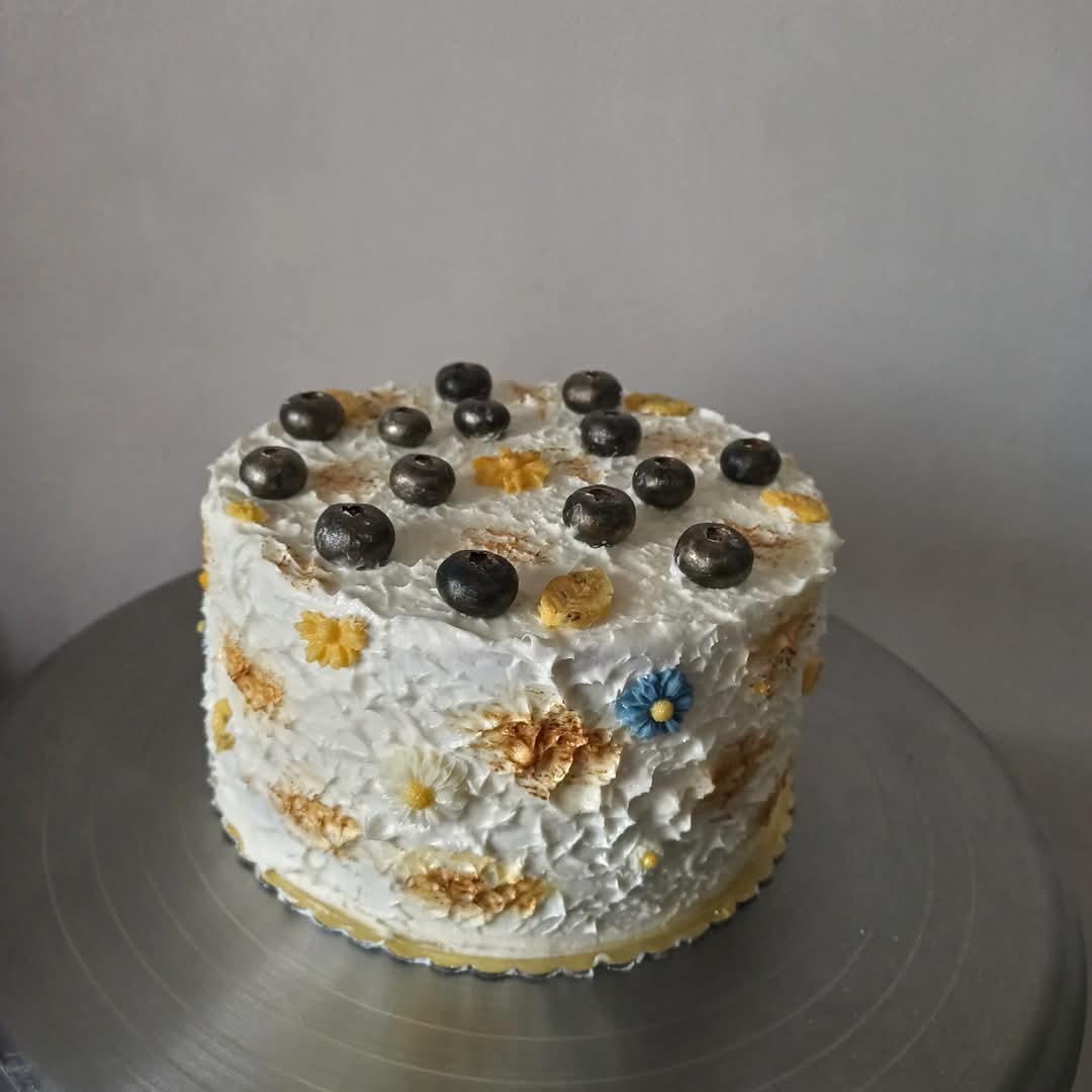 Tort zdobiony białym kremem, posypany borówkami i kwiatami. Dekoracja w stylu rustykalnym na metalowej paterze.