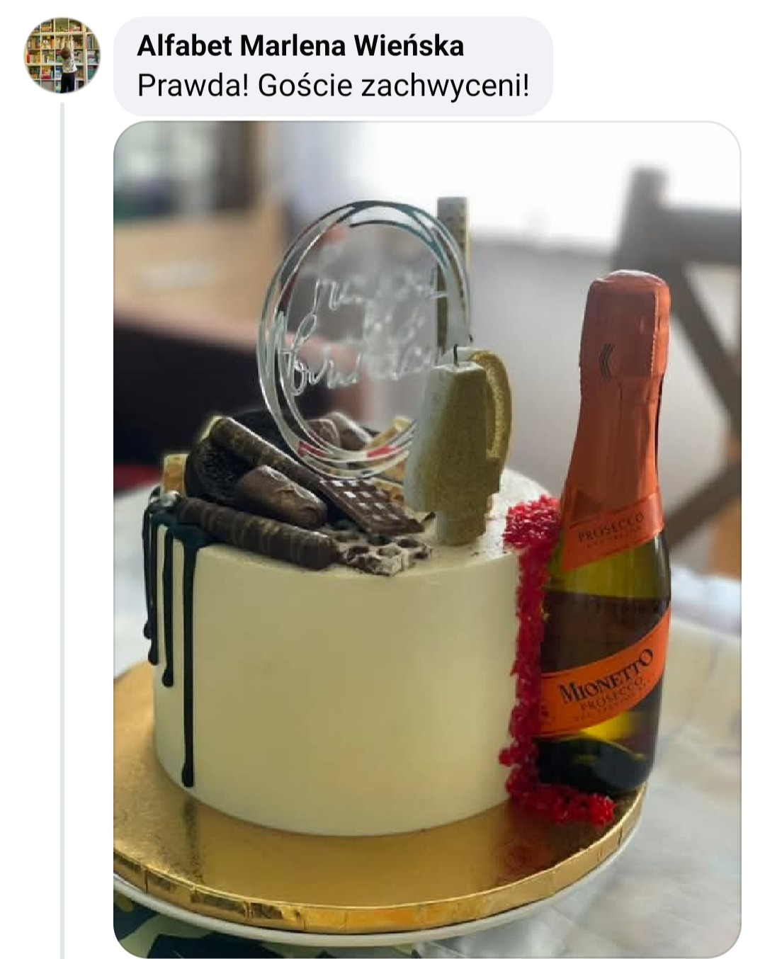 Tort urodzinowy z czekoladowymi dekoracjami, butelką Prosecco i topperem. Biały krem, złoty podkład, delikatne oświetlenie, rozmyte tło.