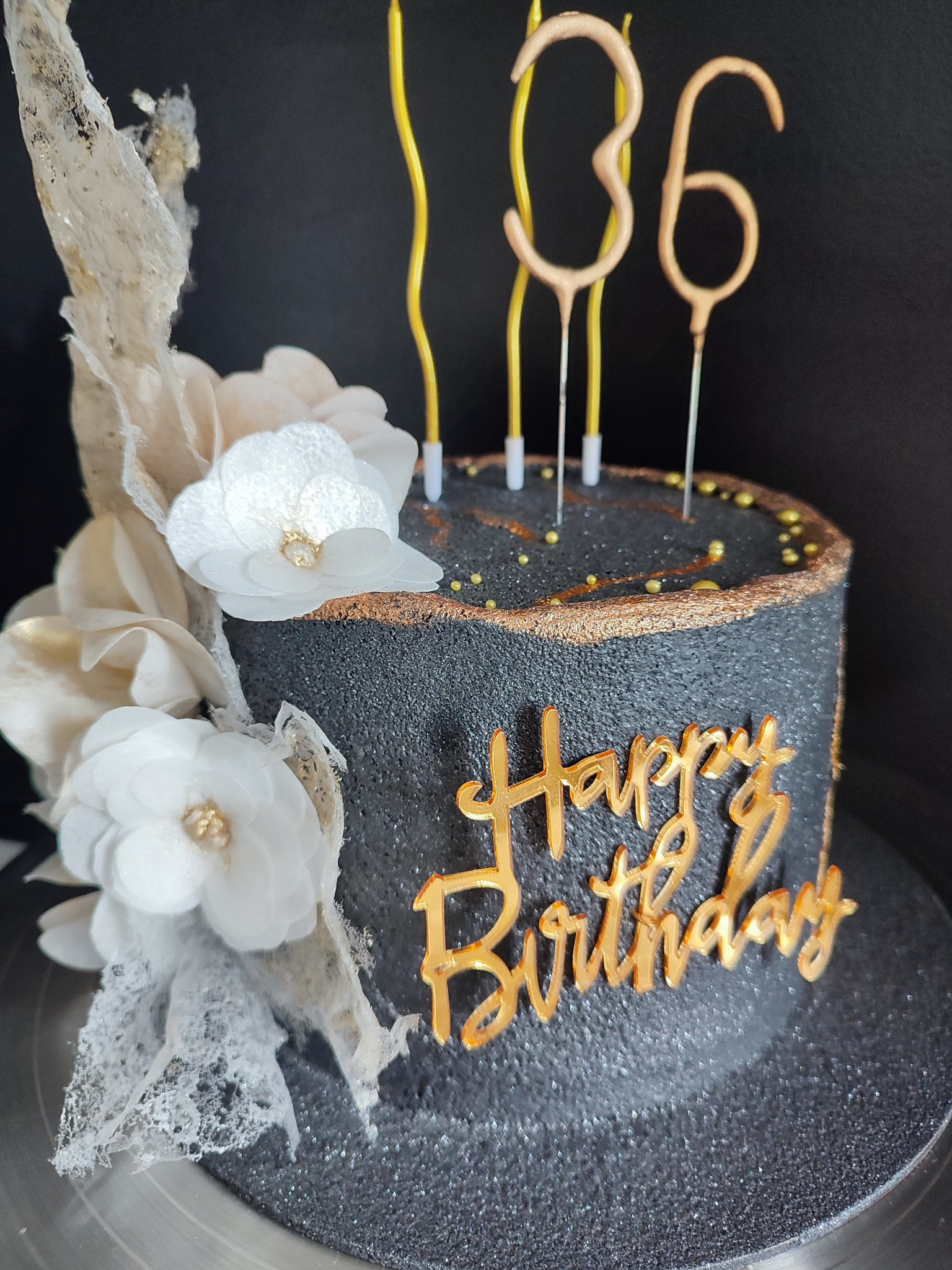 Tort urodzinowy w ciemnej tonacji z dekoracjami z białych kwiatów i napisem 'Happy Birthday' w złotym kolorze, świeczki w kształcie cyfr '36'.