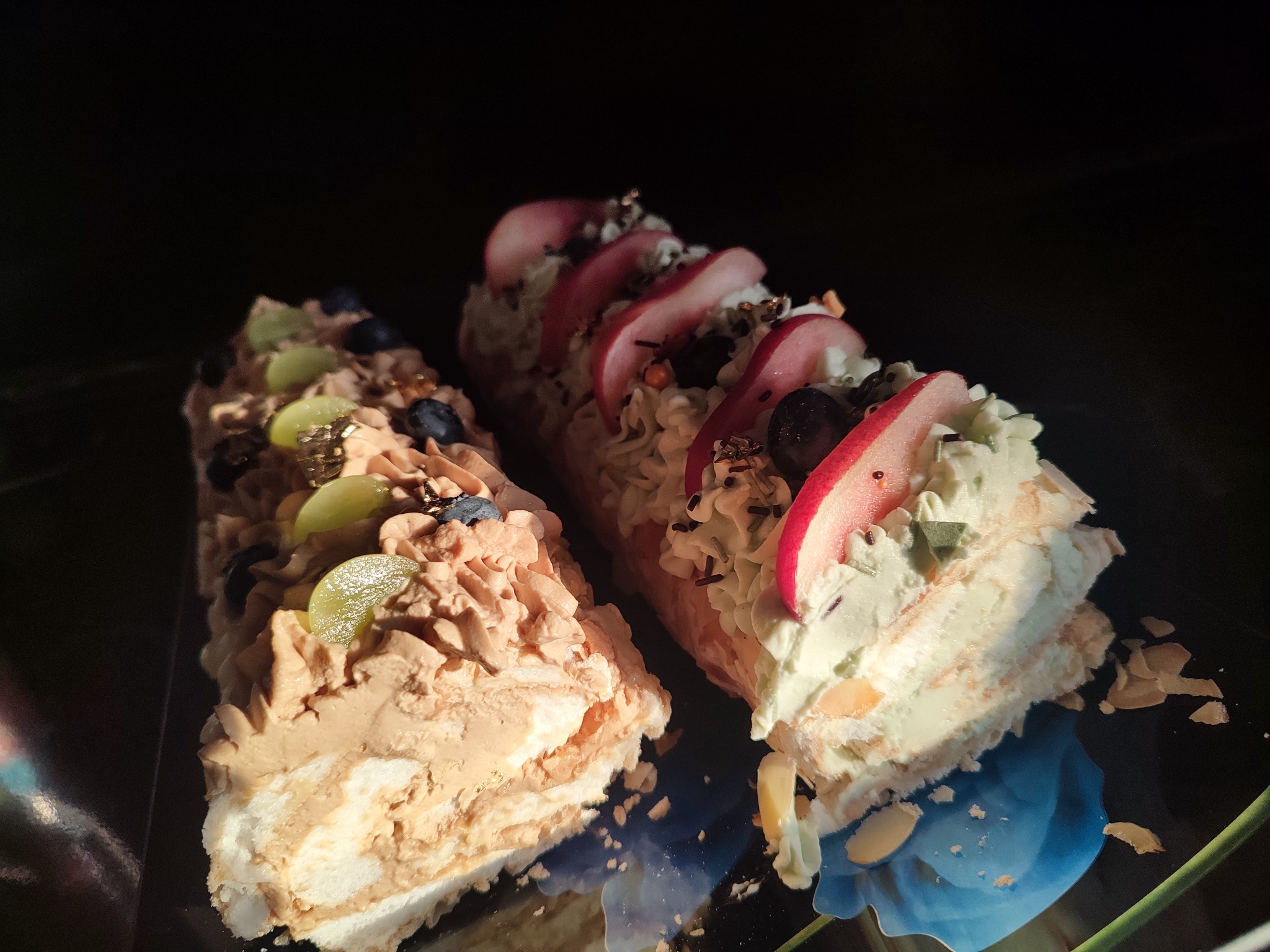 Dwa kawałki tortu bezowego na ciemnym talerzu, jeden z kremem czekoladowym i winogronami, drugi z kremem pistacjowym i brzoskwiniami, w słonecznym świetle.