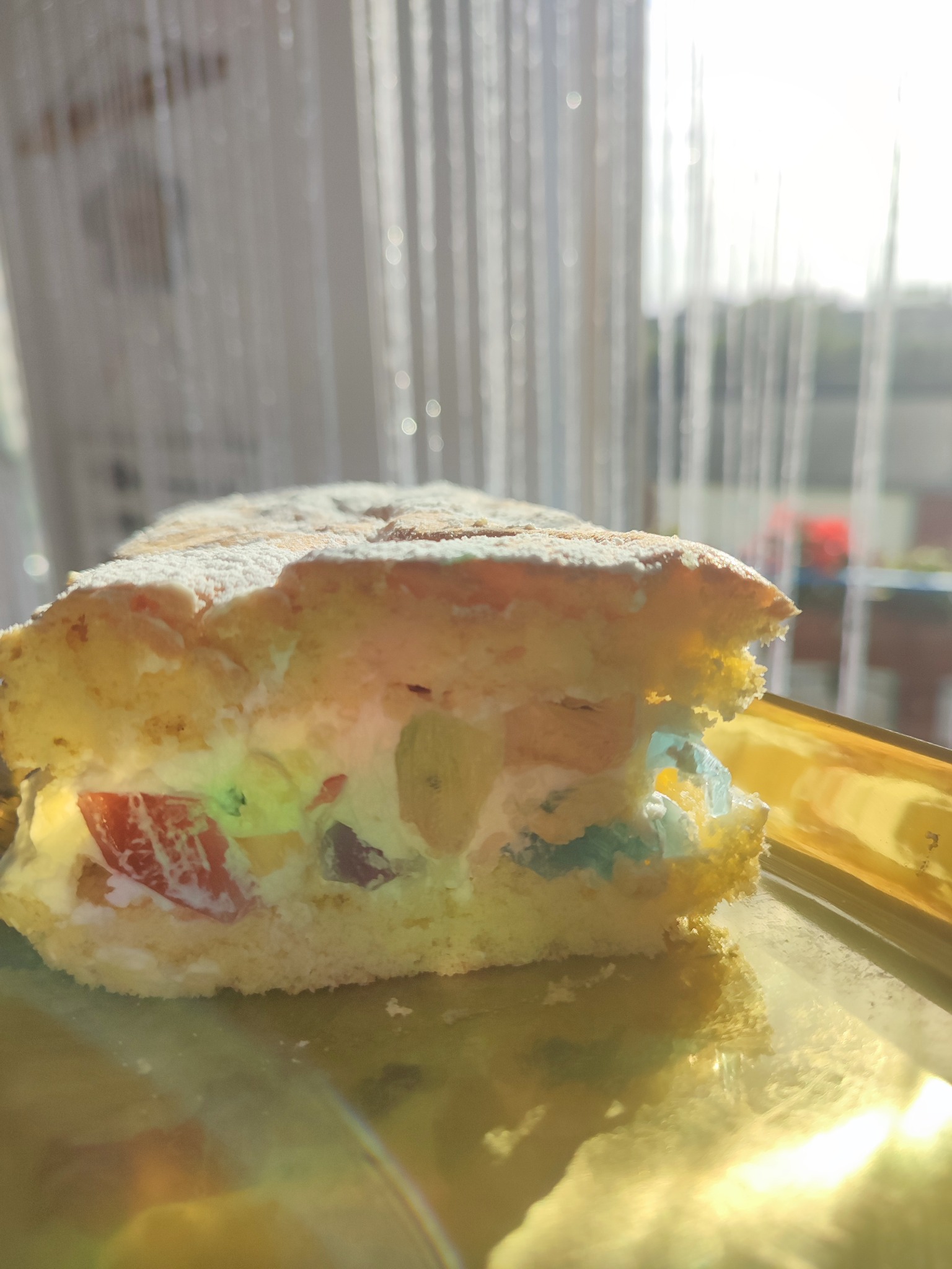 Kawałek tortu owocowego z kremem i galaretką na złotej paterze, posypany cukrem pudrem, w tle błyszcząca zasłona i okno, jasne światło.