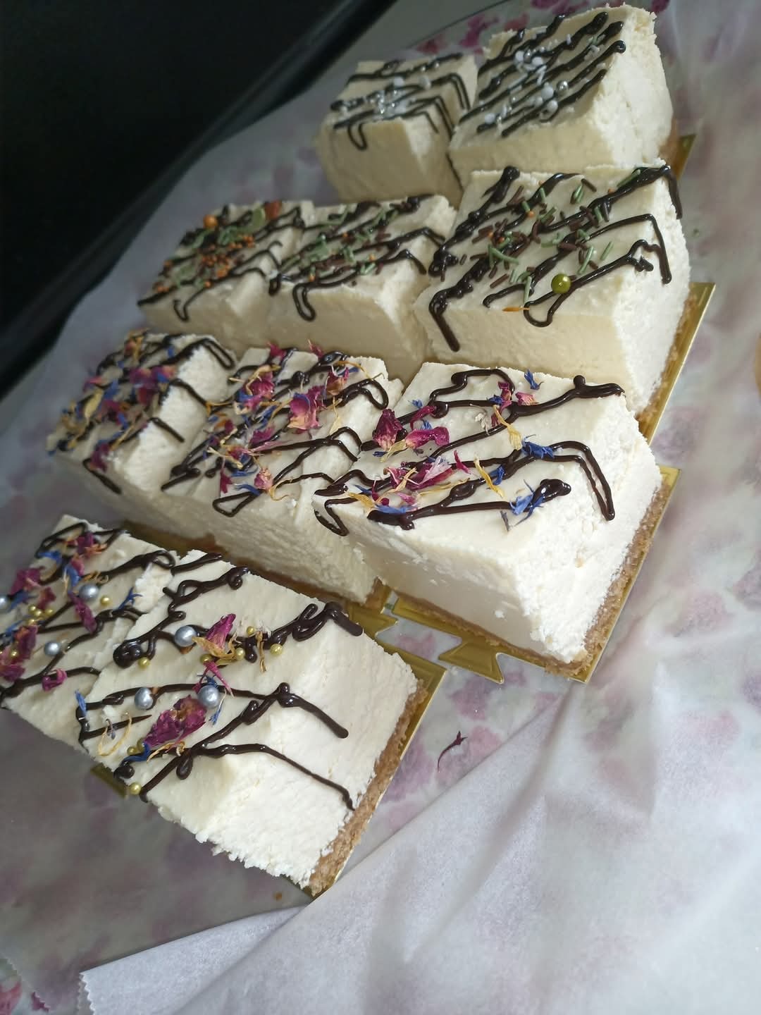 Kremowe kostki sernika udekorowane polewą czekoladową i posypką z kwiatów oraz srebrnych kulek, ułożone na papierze z motywem kwiatowym.