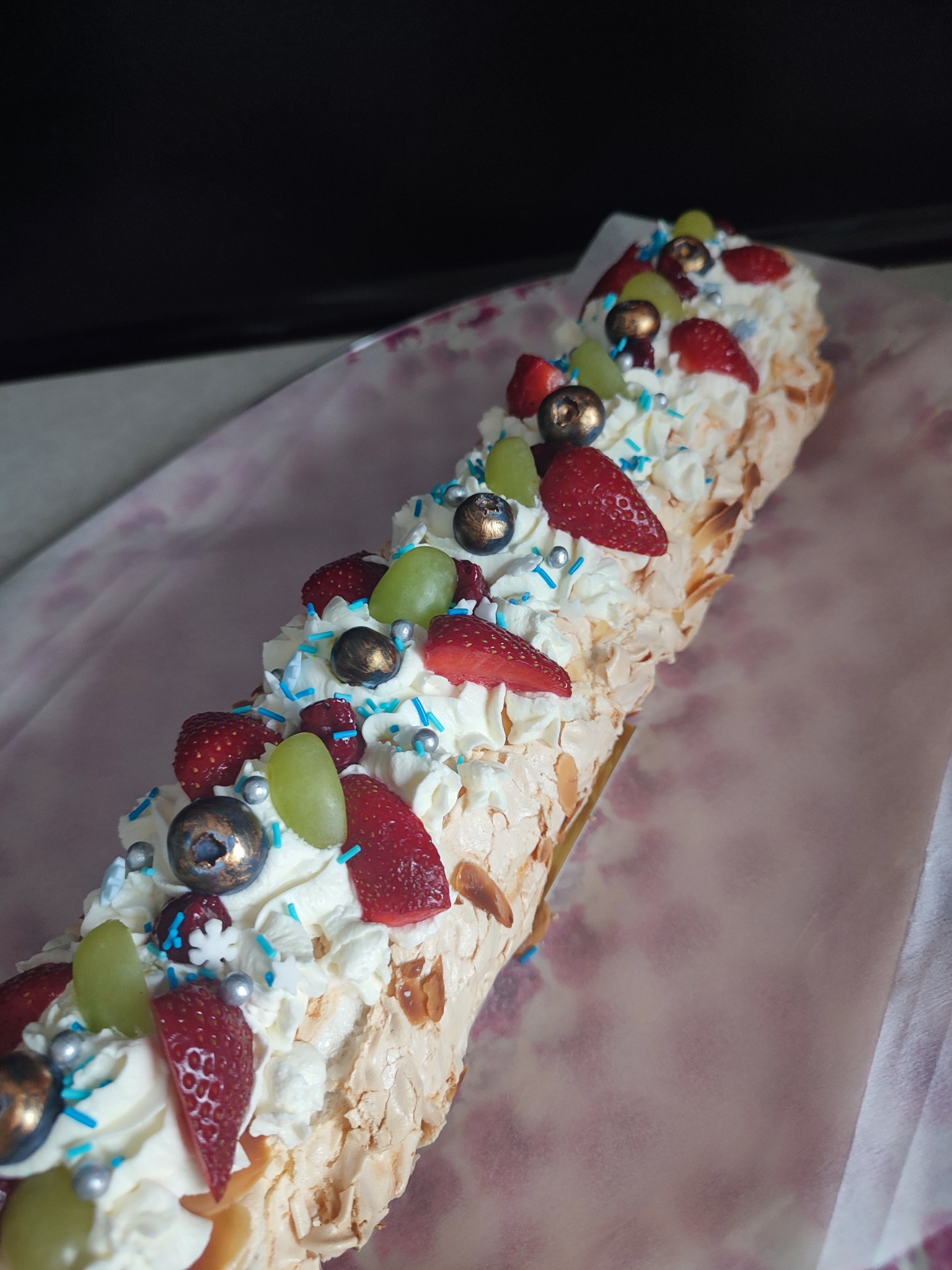 Długi tort bezowy, udekorowany bitą śmietaną, truskawkami, winogronami i srebrnymi kuleczkami, leży na papierze w delikatne kwiaty.