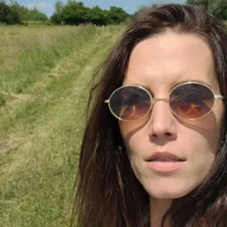 Portret kobiety w okularach przeciwsłonecznych na tle łąki z drzewami i błękitnego nieba z chmurami, wykonany w słoneczny dzień.