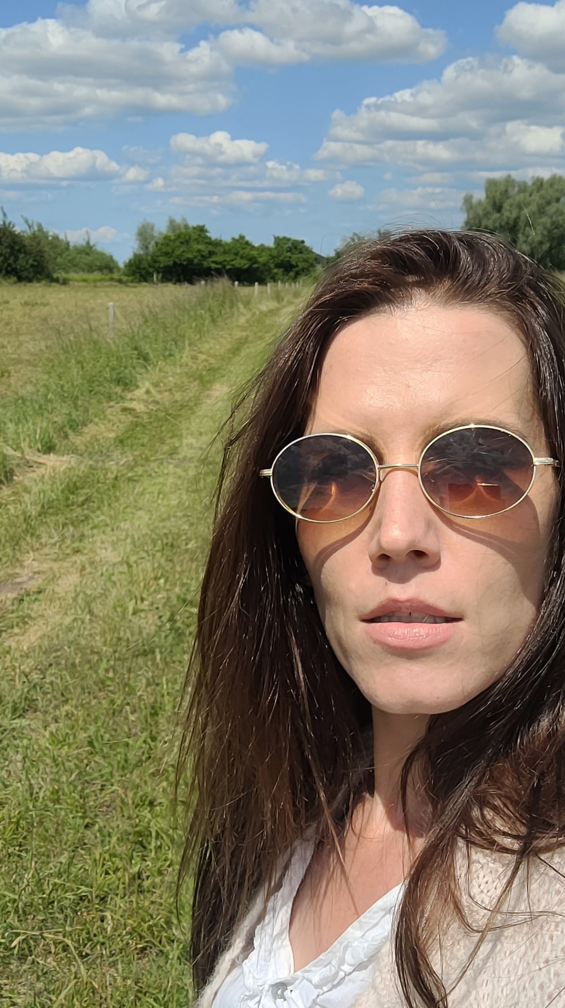 Portret kobiety w okularach przeciwsłonecznych na tle łąki z drzewami i błękitnego nieba z chmurami, wykonany w słoneczny dzień.