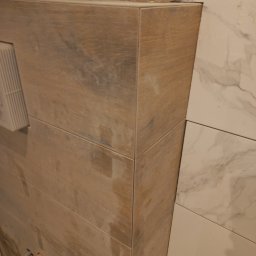 Mariusz Leonowski - Wykończona obudowa WC z płytek imitujących drewno, sąsiadująca z marmurowymi płytkami na ścianie. Widoczne elementy instalacji wodnej.