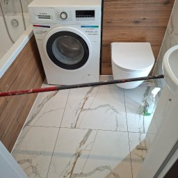 Spid bud - Białe płytki imitujące marmur w łazience z pralką Bosch, toaletą i umywalką. Widoczny fragment kija malarskiego i butelka ze środkiem czyszczącym.
