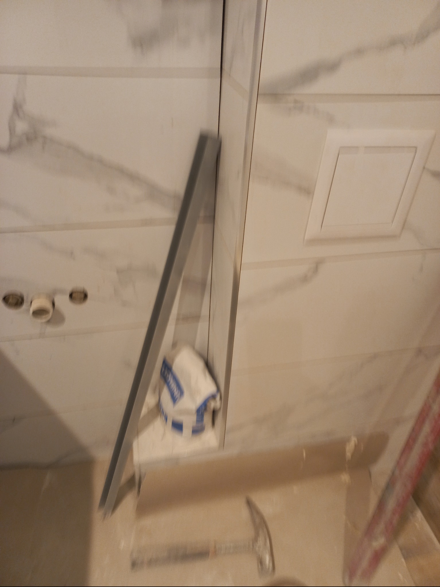 Białe płytki imitujące marmur na ścianie łazienki, widoczny odpływ liniowy, worek z klejem i młotek. Etap prac wykończeniowych.