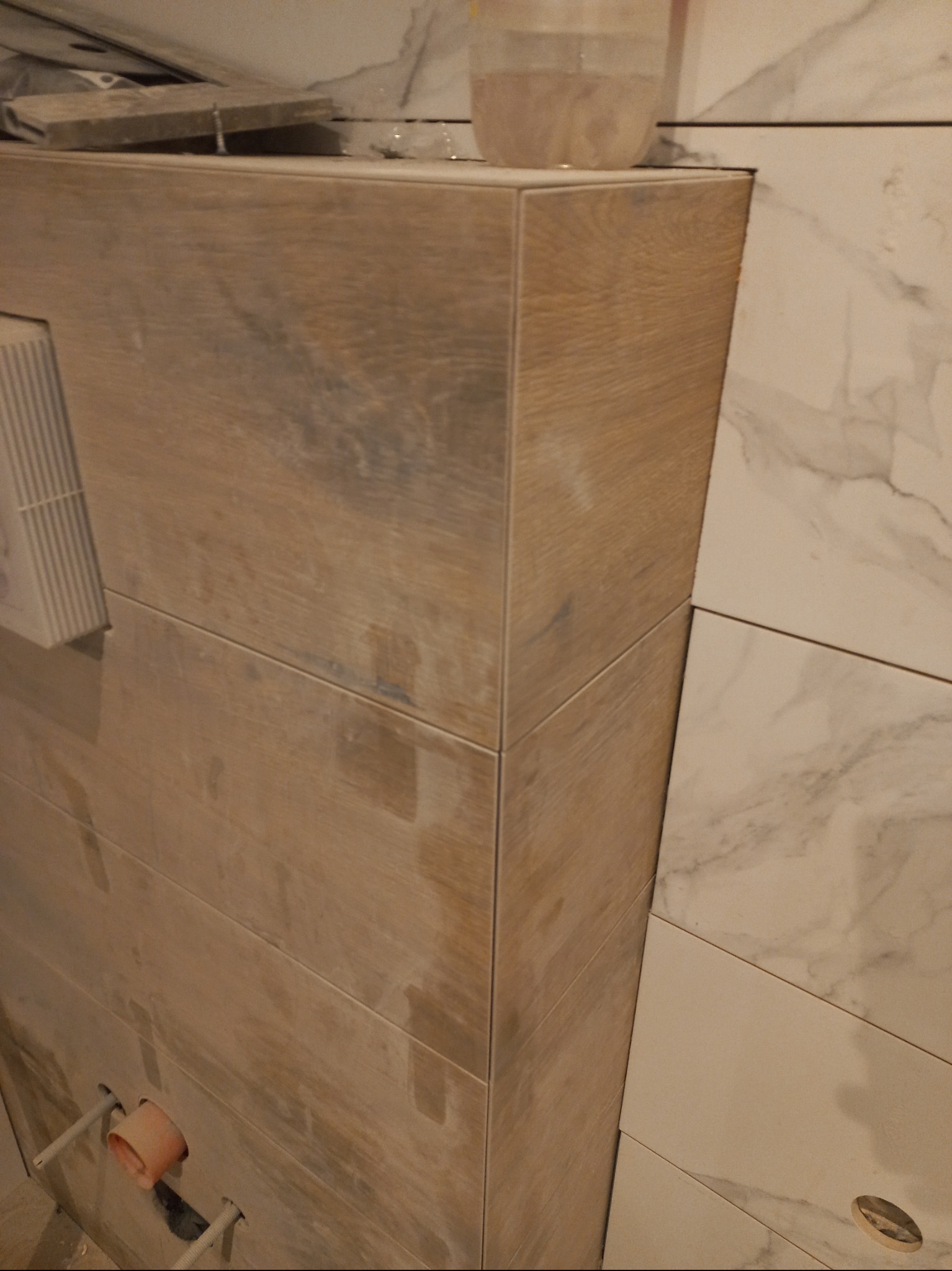 Wykończona obudowa WC z płytek imitujących drewno, sąsiadująca z marmurowymi płytkami na ścianie. Widoczne elementy instalacji wodnej.