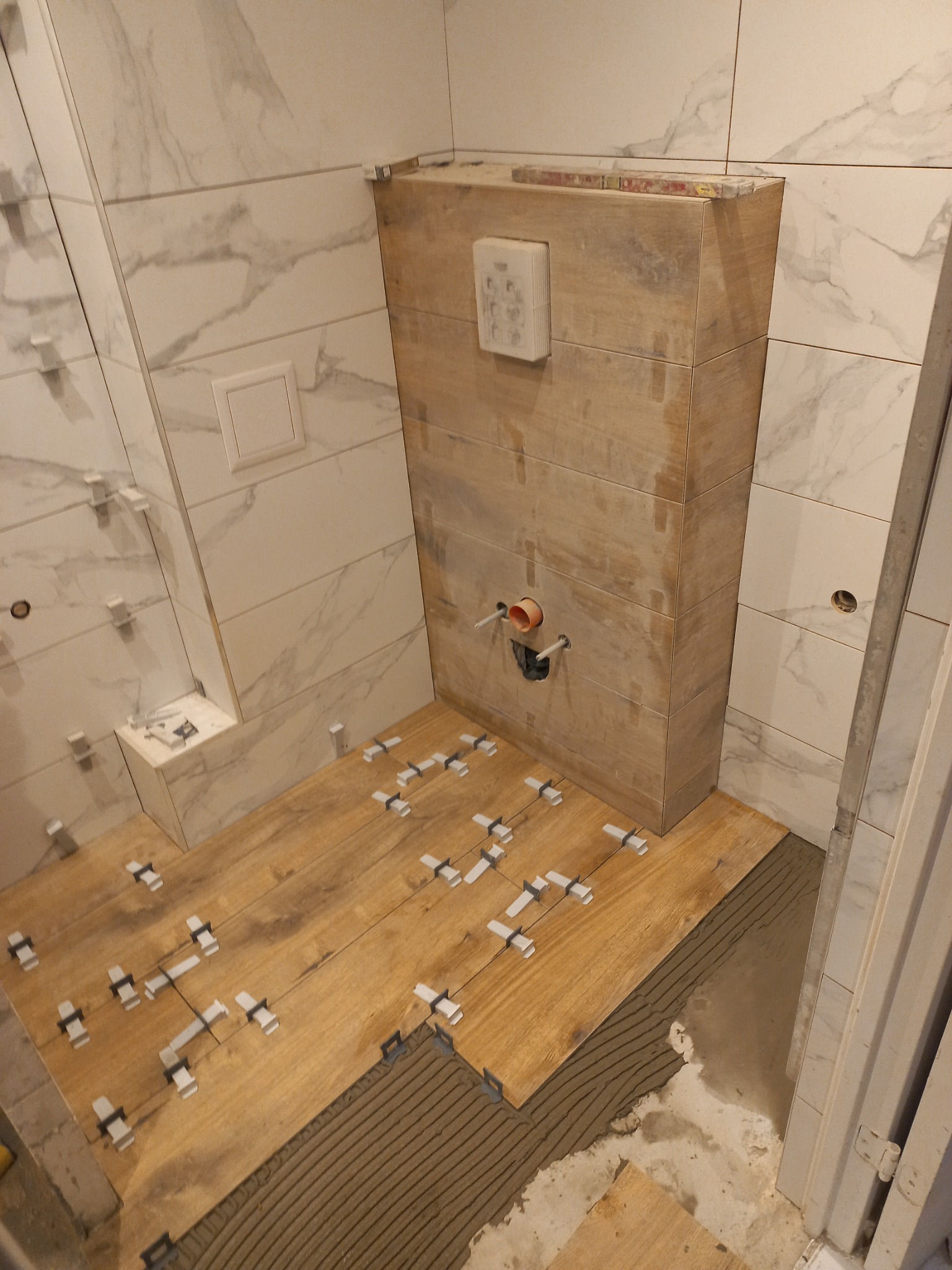 Łazienka w trakcie wykańczania: drewnopodobne płytki podłogowe z krzyżykami dystansowymi, marmurowe płytki ścienne i zabudowa stelaża WC. Widoczne etapy prac.