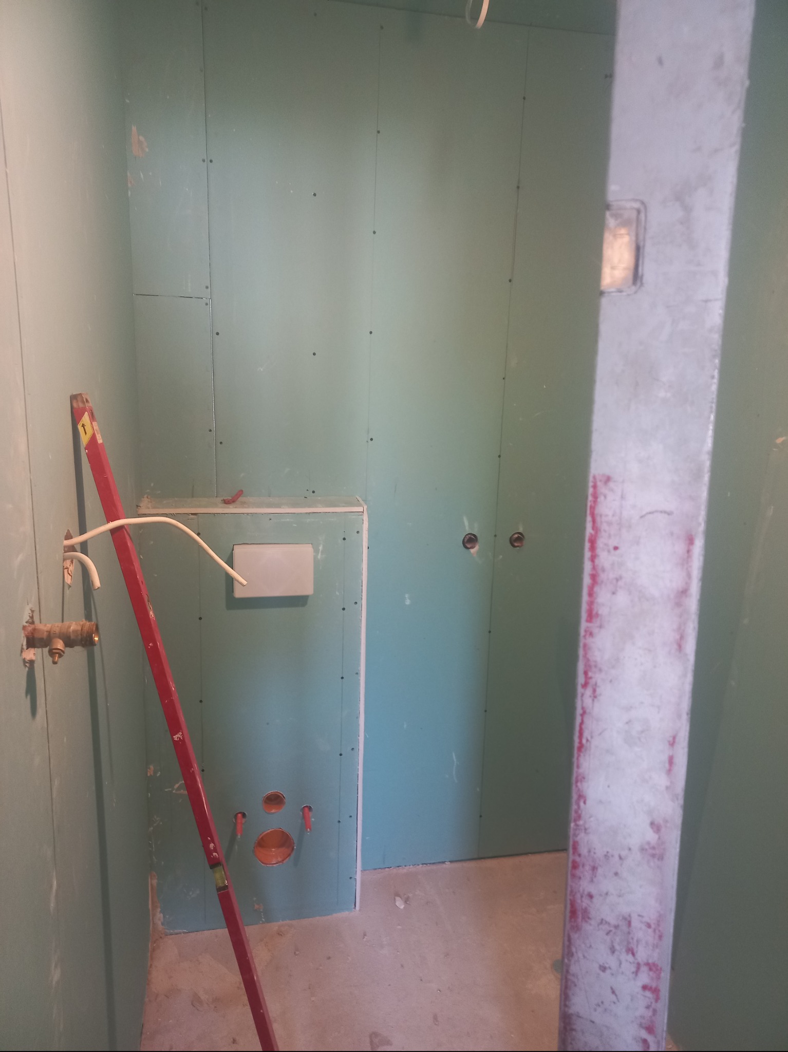 Łazienka w trakcie montażu płyt karton-gips, widoczne instalacje wodne i elektryczne, poziomica oparta o ścianę. Surowy stan wykończenia, przygotowanie do dalszych prac.