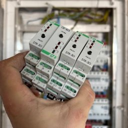 ACI PIOTR ZIELEŃ - Ręka trzymająca cztery białe moduły STR-3D z zielonymi akcentami, na tle otwartej skrzynki elektrycznej z widocznymi przewodami i elementami instalacji.