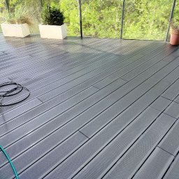 Czysta Przestrzeń - Ciemnoszara, mokra deska tarasowa po czyszczeniu, z widocznym wężem i dyszą myjki ciśnieniowej, w tle zieleń. Widok na taras po umyciu.