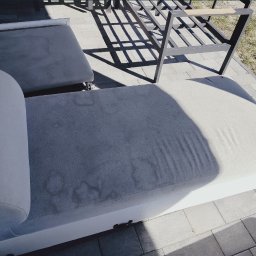 Czysta Przestrzeń - Z bliska: plamy na jasnej tapicerce sofy ogrodowej, widoczne na tle kostki brukowej i cienia metalowej ramy meblowej. Perspektywa z góry.