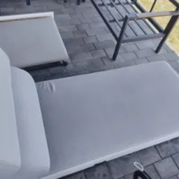 Czyszczenie jasnej, tapicerowanej leżanki na zewnątrz, widoczny odkurzacz piorący na kostce brukowej. Metalowa konstrukcja leżanki w tle.