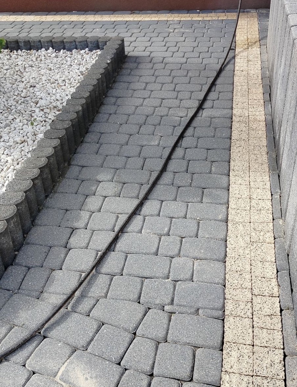 Szara kostka brukowa z obrzeżami i kablem na powierzchni. Ujęcie z góry, ukazujące detale tekstury i kontrast kolorów między kostką a obrzeżami.