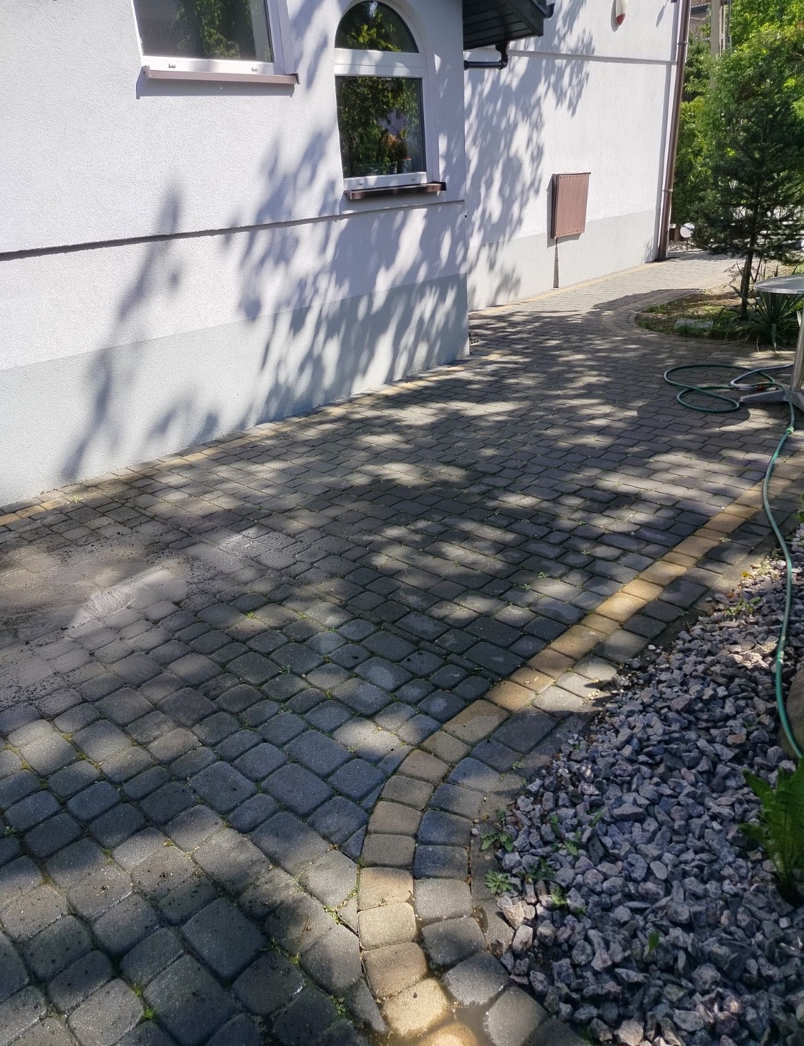 Częściowo umyta kostka brukowa przed domem, z widocznym kontrastem czystości. W tle biała elewacja budynku i zieleń drzew. Wąż ogrodowy leży obok.