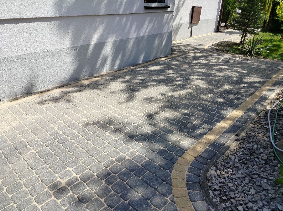 Ułożona kostka brukowa w odcieniach szarości i beżu, z widocznymi cieniami drzew, obok ściany budynku. Estetyczne wykończenie obrzeży i dbałość o detale.
