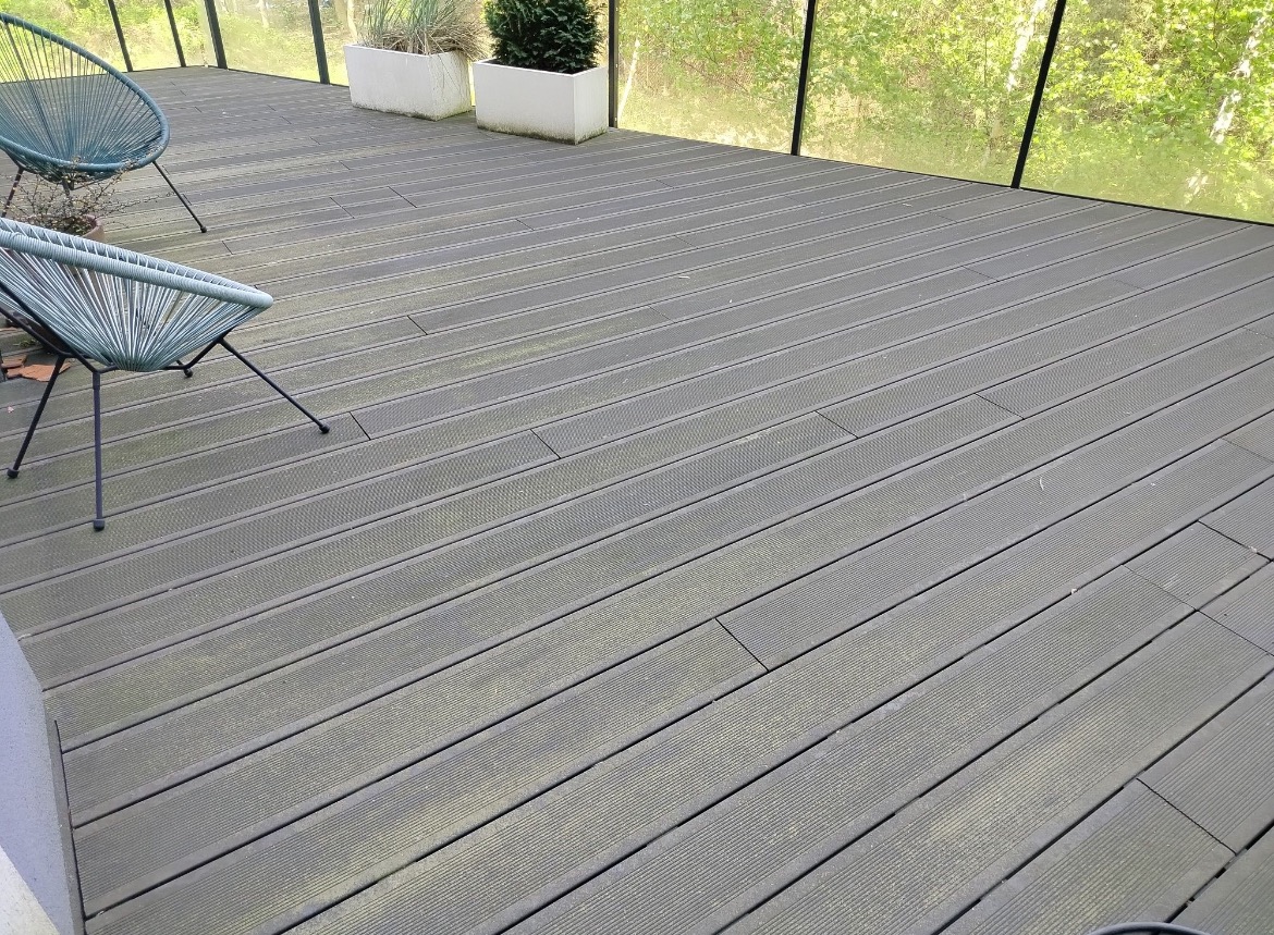 Szara deska tarasowa z widocznymi zabrudzeniami, dwa krzesła z plecionki i donice z roślinami w tle, przed myciem. Układ desek wzdłuż kadru, lekki kąt.