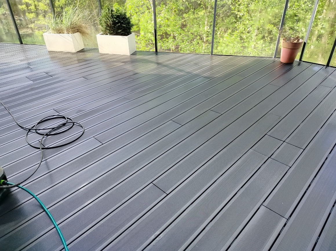 Ciemnoszara, mokra deska tarasowa po czyszczeniu, z widocznym wężem i dyszą myjki ciśnieniowej, w tle zieleń. Widok na taras po umyciu.
