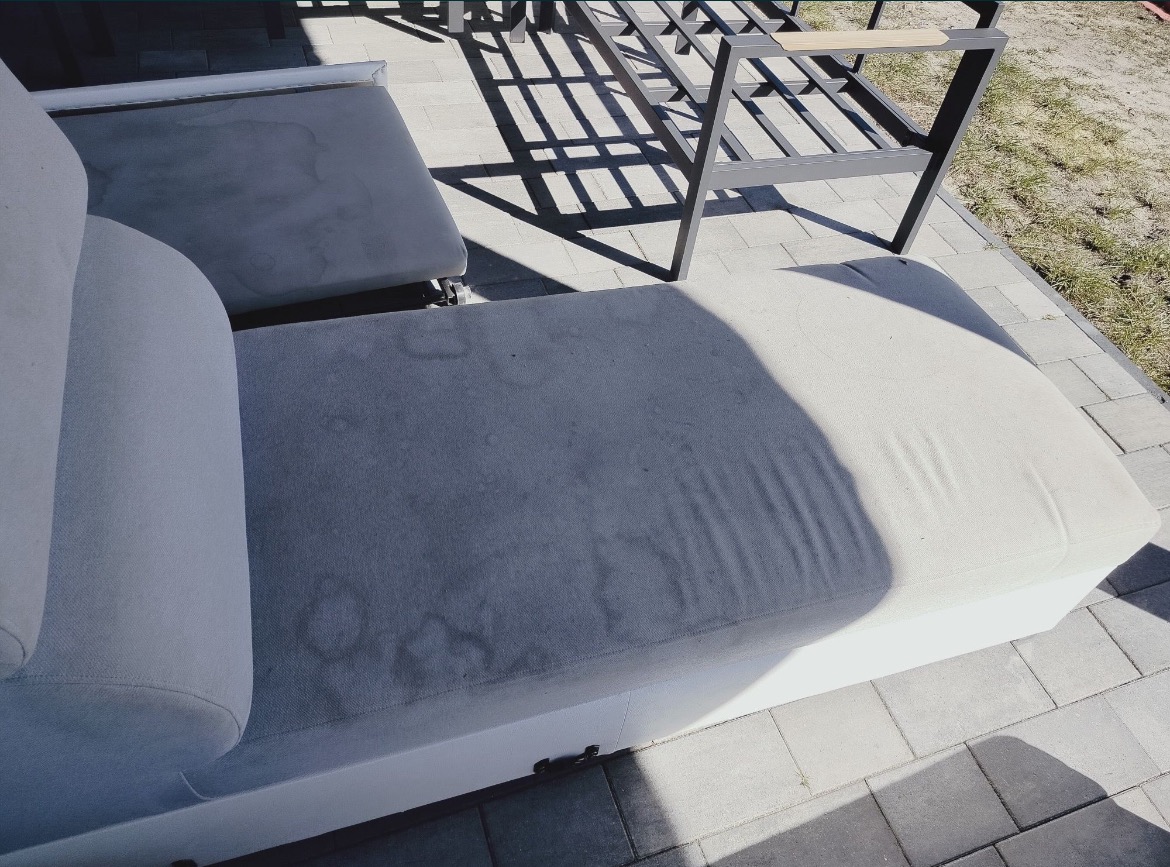 Z bliska: plamy na jasnej tapicerce sofy ogrodowej, widoczne na tle kostki brukowej i cienia metalowej ramy meblowej. Perspektywa z góry.