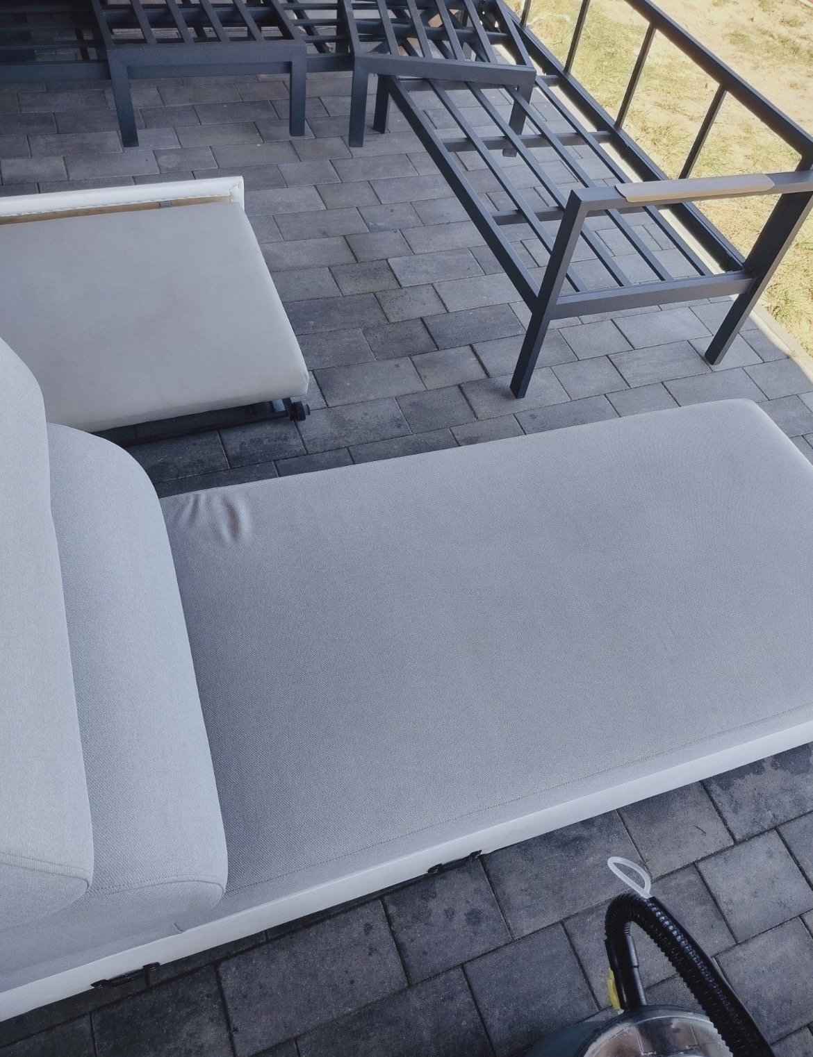 Czyszczenie jasnej, tapicerowanej leżanki na zewnątrz, widoczny odkurzacz piorący na kostce brukowej. Metalowa konstrukcja leżanki w tle.