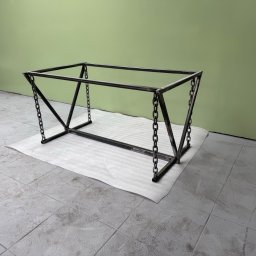 DAN-TIG GROUP SPÓŁKA Z OGRANICZONĄ ODPOWIEDZIALNOŚCIĄ - Metalowa konstrukcja stołu z łańcuchami, spawana, surowa, na białej piance. Nowoczesny design, zielona ściana w tle. Elementy spawane pod kątem.